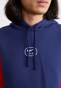 Hoodie bleu marine avec un cordon de serrage, présentant un logo blanc sur la poitrine. Le matériau semble doux, avec des manches raglan et une texture lisse.
