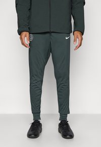 Grønne treningbukser med smale ben, laget av glatt stoff, med Nike-logo og et sirkulært emblem. Sammen med svarte treningstøvler.