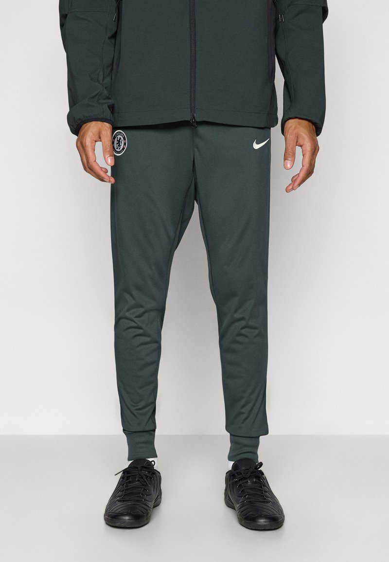 Grønne treningbukser med smale ben, laget av glatt stoff, med Nike-logo og et sirkulært emblem. Sammen med svarte treningstøvler.