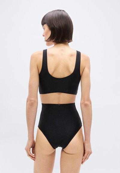 Γυναίκα με κοντά καστανά μαλλιά που φοράει μαύρο αμάνικο crop top και μαύρα μαγιό με ψηλή μέση, γυρισμένη από την πλάτη, πάνω σε απλό φωτεινό φόντο.