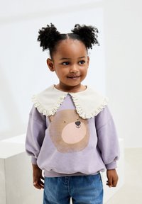 Felpa viola con grafica di un orso e colletto arricciato color crema. Vestibilità comoda con maniche a sbuffo e polsini a coste. Jeans in denim blu.