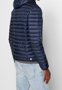 Marinblå pufferjacka med huva, med quiltat mönster och glänsande yta. Inkluderar en liten logotypdetalj nära fållen.