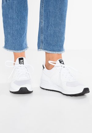 Person iført hvide New Balance 574 sneakers og blå jeans med frynset kant, stående på en hvid gulv mod en hvid baggrund.