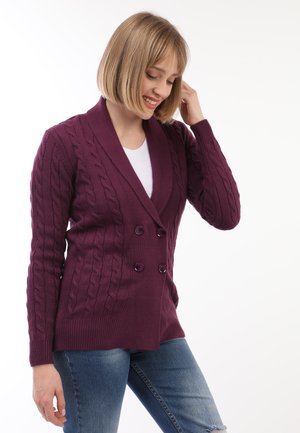 Doppelt geschnittene Strickjacke in tiefem lila Zopfmuster. Mit großen schwarzen Knöpfen und einem Schalkragen. Kombiniert mit hellblauen Jeans.