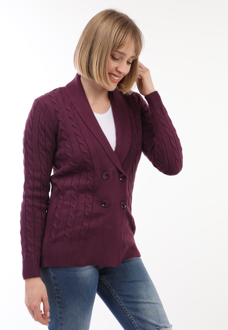 Doppelt geschnittene Strickjacke in tiefem lila Zopfmuster. Mit großen schwarzen Knöpfen und einem Schalkragen. Kombiniert mit hellblauen Jeans.