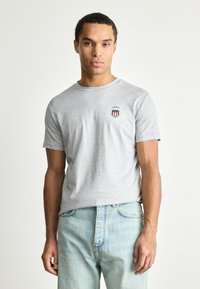 Grå bomulds t-shirt med korte ærmer og rund halsudskæring; har et cirkulært logo i rød og blå på venstre brystområde.