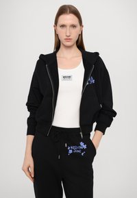 Musta vetoketjullinen huppari, jossa on suuret taskut ja kukka-embroidery. Yhdistetty valkoiseen toppiin ja mustiin verkkareihin, joissa on vastaavat kukkakoristeet.