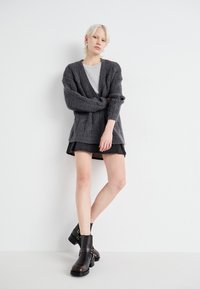 Grauer Strickcardigan mit V-Ausschnitt, strukturiertem Muster, langen Ärmeln, kombiniert mit einem hellgrauen Hemd und einem schwarzen Minirock, akzentuiert durch schwarze Stiefel.