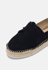 Espadrille noire à enfiler avec dessus en tissu noué et semelle en jute tressée sur fond blanc.