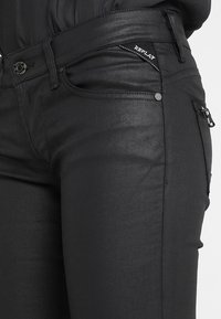 Vaqueros de denim negros con un diseño elegante, detalle de bolsillo con cremallera y etiqueta de marca en la cintura, con una textura suave y ligeramente brillante.