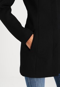 Černý vlněný kabát s elegantním designem a bočními kapsami, hladkou texturou, doplněný o světle modré džínové kalhoty.