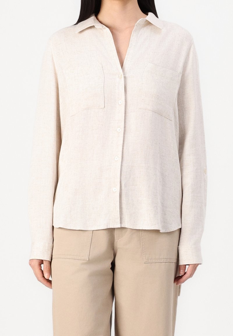 Femme portant une chemise beige clair à manches longues avec deux poches poitrine et un pantalon beige sur un fond uni.