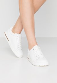 Sneakers bianchi in pelle con punta rotonda, pannelli laterali testurizzati e suola piatta in gomma. Presentano lacci bianchi e un discreto accentuazione marrone sul lato.
