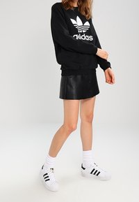Person i svart Adidas-tröja, svart läderminikjol, vita strumpor och vita Adidas-sneakers med svarta ränder står på en vit yta.