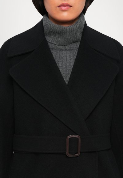Femme portant un manteau en laine noir ceinturé sur un pull en torsades gris à col roulé, mettant en valeur le col et le haut du torse.