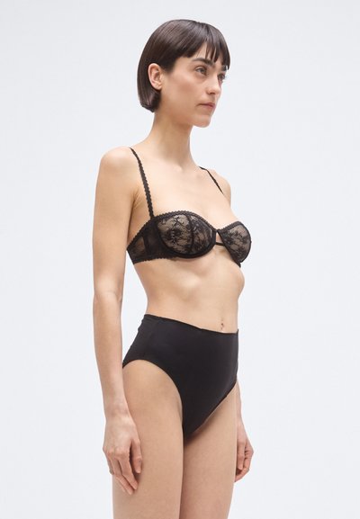 Melns mežģīņu krūšturis ar stīpām un regulējamām lencītēm, savienots ar augstām melnām biksītēm ar gludu, matētu virsmu.