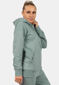 Felpa verde chiaro in tessuto testurizzato, con tasca anteriore, cappuccio con cordini e polsini a costine, indossata con pantaloni abbinati.