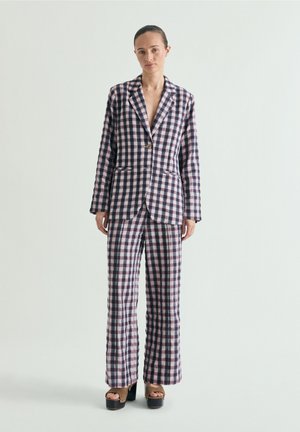 Mujer de pie con una blazer de cuadros a juego y pantalones de pierna ancha con sandalias de plataforma sobre un fondo claro liso.