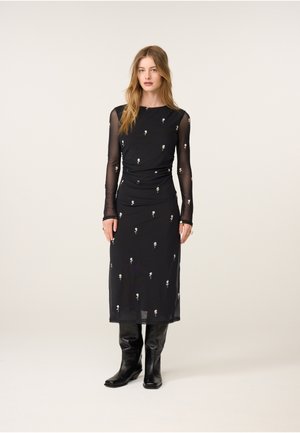 Frau trägt ein schwarzes transparentes Midikleid mit langen Ärmeln und kleinen weißen Blumenmustern, kombiniert mit schwarzen knielangen Stiefeln.