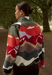 Giacca in pile con un vivace motivo multicolore di montagne, caratterizzata da tonalità di rosso, verde e grigio; colletto alto e tessuto testurizzato.