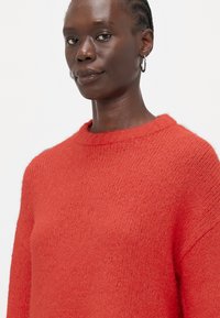 Pullover rosso in maglia con scollatura a girocollo e maniche lunghe. Tessuto morbido e testurizzato con una vestibilità leggermente over. Finitura con un colletto a costine.