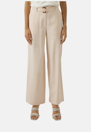 Femme portant un pantalon large taille haute beige avec de grandes poches avant, une ceinture assortie et des sandales grises à brides torsadées.