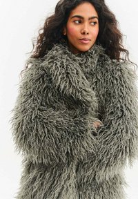 Kobieta o długich kręconych włosach, ubrana w gruby, szary, puszysty płaszcz ze sztucznego futra, ze skrzyżowanymi ramionami, patrząca na bok na jednolitym białym tle.