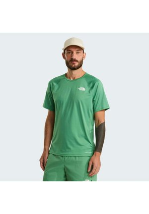 Zöld rövid ujjú ingben és rövidnadrágban álló férfi, fehér logóval, bézs sapkával és sötét tetoválással a bal alkarján, világos háttér előtt.