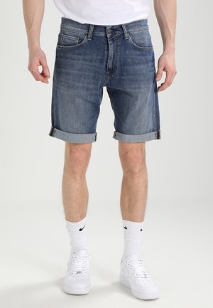 Mann trägt hochgekrempelte blaue Jeansshorts, weiße Socken und weiße Turnschuhe und steht vor einem einfarbigen Hintergrund.