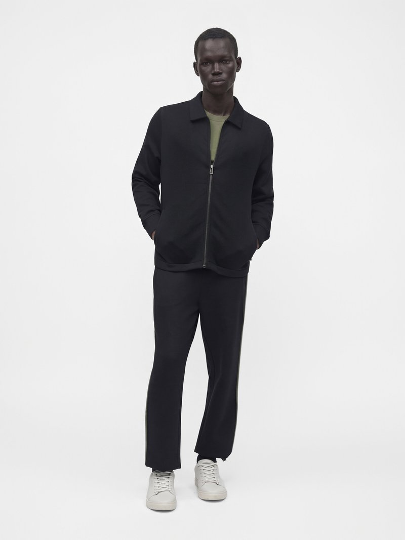 Homme debout les mains dans les poches, portant une veste zippée noire, un pantalon noir, une chemise vert olive et des baskets blanches sur un fond uni.