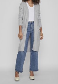 Grå stickad cardigan med långa ärmar, öppen front och fickor, kombinerad med en vit V-ringad skjorta och vida blå jeans.