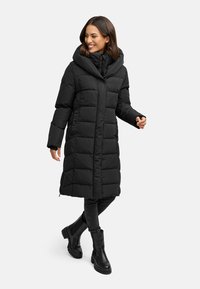 Schwarzer gepolsterter Mantel mit Kapuze, Knopfverschluss, gesteppten Abschnitten und Seitentaschen. Kombiniert mit schwarzen Skinny Jeans und Stiefeletten.