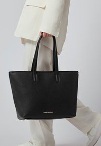 Persona in abito beige che tiene una grande borsa a tracolla nera in pelle con doppi manici e il logo dorato "Victoria Hyde London".