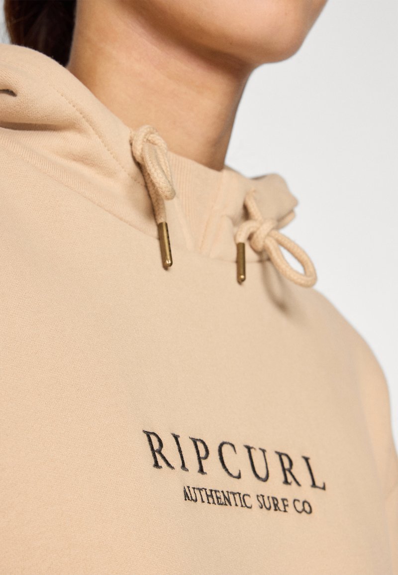 Beżowa bluza z kapturem z regulowanymi sznurkami i złotymi metalowymi akcentami. Na przodzie znajduje się czarny logotyp "RIPCURL AUTHENTIC SURF CO".
