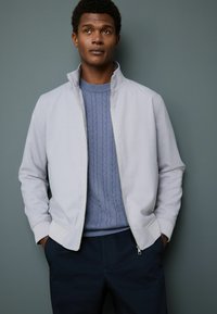 Young man with hands in pockets wearing light gray zip-up jacket over blue cable-knit sweater against a gray background.
