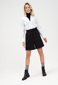 Camicia bianca con colletto, pantaloni corti neri a vita alta e stivaletti neri con lacci alla caviglia. Calzini grigi visibili. Design semplice, sartoriale e con una vestibilità rilassata.