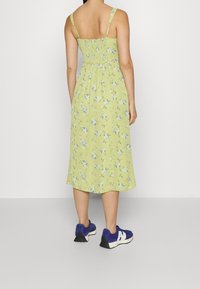 Femme portant une robe midi à fleurs vert clair avec des bretelles fines et des baskets violettes, se tenant devant un fond blanc uni.