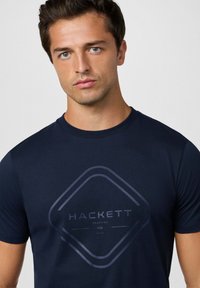 Marinblå t-shirt i mjukt tyg, med en mörkgrå diamantlogotyp och texten "HACKETT" i slim fit. Rund hals.