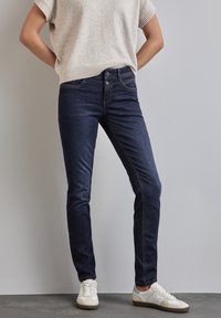 Mörkblå skinny jeans i denim, med knappslutning fram och kontrastsömmar. Matchas med en ljusbeige stickad topp och vita sneakers.