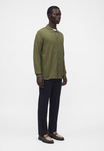 Homme debout portant une chemise à manches longues boutonnée vert olive, un pantalon noir et des mocassins marron et beige sur un fond blanc uni.