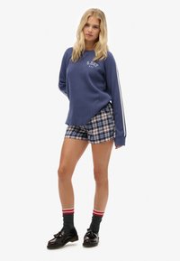 Sudadera azul con relieve y rayas blancas en las mangas, combinada con pantalones cortos de cuadros en azul, rojo y blanco. Zapatos negros con acentos de lazo y calcetines grises.