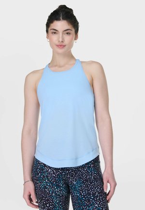 Vrouw met donker haar in een blauw mouwloos topje en zwart-blauw-witte legging met patroon, staand tegen een effen achtergrond.