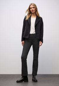 Giacca nera cropped con ampi rever, abbinata a una maglietta bianca e jeans scuri. Il modello indossa stivaletti neri alla caviglia. Design semplice e su misura.