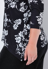 Nahaufnahme einer Person, die ein schwarzes Oberteil mit weißen Blumenmustern und schwarze gerippte Hosen trägt, die Hand entspannt an ihrer Seite gegen eine helle Wand.