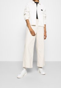 Krämfärgad kordjacka, svart topp och vida beige byxor, kombinerat med vita sneakers. Noterbara tatueringar och ringar på handen.