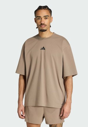 Mand med flettet hår iført en løs beige Adidas t-shirt og matchende shorts, stående mod en ensfarvet lys baggrund.