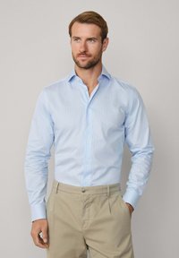 Man in een lichtblauw overhemd, gestopt in een beige broek, staat met één hand in zijn zak tegen een plain achtergrond.