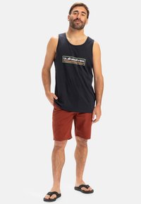Svart ärmlös tanktop med en färgglad logotyp, kombinerad med rostfärgade shorts och svarta flip-flops. Lätt och ledig strandstil.