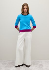 Maglione azzurro chiaro con rifiniture viola e scollo nero, abbinato a pantaloni bianchi a gamba larga e calzature scure su uno sfondo neutro.