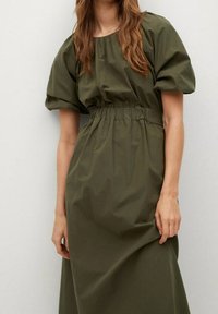 Olivengrünes Kleid mit puffärmel, elastischem Bund und einem fließenden Rock aus glattem Stoff, der ein schlichtes, minimalistisches Design aufweist.